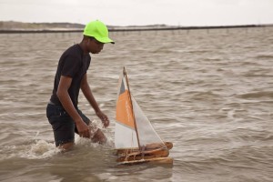 Foto de um jovem negro de boné verde brinca com um barquinho a vela de brinquedo no mar.