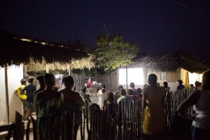 Foto ilustrando várias pessoas fora de casa, observam um telão.