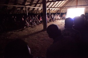 Foto de várias silhuetas de crianças observam um telão, ao fundo há mais crianças sentadas fazendo o mesmo, todos dentro de uma casa de palha.