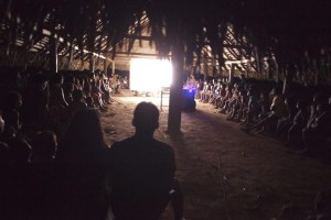 Foto com a silhueta de um homem e de uma mulher, os dois estão de costa para a fotografia e ao redor deles várias crianças sentadas dentro de uma casa de palha, todos observam um telão.