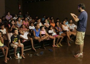 Foto de um salão onde crianças prestam atenção em um jovem de barba falando.