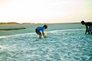 Foto de dois garotos brincando dentro da agua, com barco a vela de brinquedo, ao entardecer.