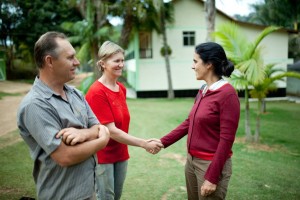 Foto composta por três pessoas interagindo, duas mulheres estão se cumprimentando, enquanto um homem as observa.
