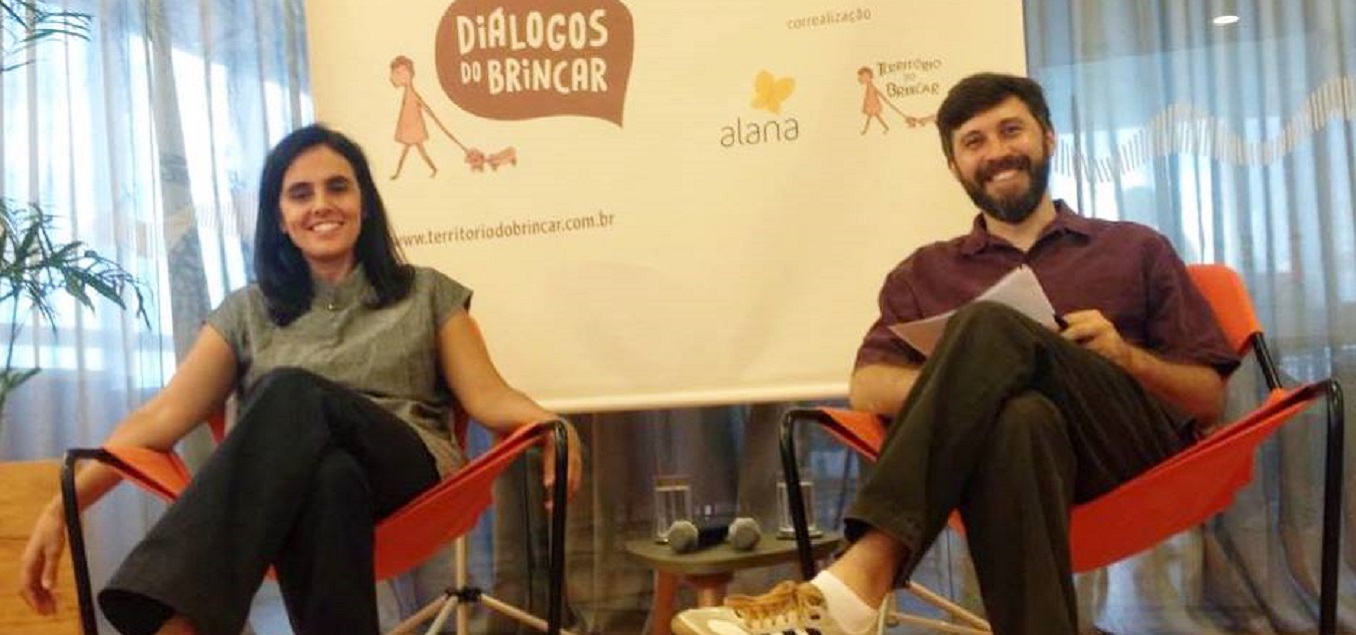 Foto com duas pessoas sentadas, uma mulher e ao seu lado esquerdo um homem de barba, ambos estão de pernas cruzadas e sorrindo.