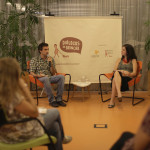 Foto composta por várias pessoas sentadas em círculo, ao fundo um homem fala ao microfone e a sua esquerda uma mulher o observa.