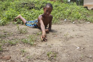Foto de um garoto negro deita em um chão de terra, com seu braço direito prepara para arremessar uma bolinha de gude.