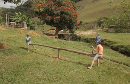 Foto com três pessoas, duas crianças e um homem, as duas crianças estão sentadas em uma gangorra feita com um tronco de uma arvore, enquanto ao fundo um home as observa.