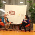 Foto com duas pessoas, uma idosa de cabelos grisalhos que fala ao microfone, ao seu lado esquerdo uma mulher observa a senhora.