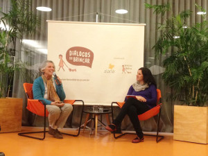 Foto com duas pessoas, uma idosa de cabelos grisalhos que fala ao microfone, ao seu lado esquerdo uma mulher observa a senhora.