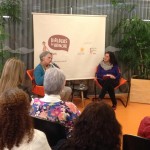 Foto composta por várias pessoas sentadas que observam duas pessoas sentadas ao fundo, uma das duas pessoas é uma idosa de cabelos grisalhos fala ao microfone, enquanto a sua esquerda a segunda pessoas que é uma mulher a observa.