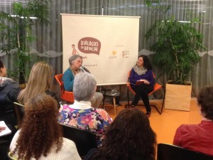 Foto composta por várias pessoas sentadas que observam duas pessoas sentadas ao fundo, uma das duas pessoas é uma idosa de cabelos grisalhos fala ao microfone, enquanto a sua esquerda a segunda pessoas que é uma mulher a observa.