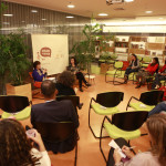 Foto composta por várias pessoas sentadas que observam ao fundo duas pessoas sentadas, uma delas é uma senhora que fala ao microfone, ao lado esquerdo dela tem uma mulher que observa a senhora.