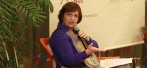 Foto de uma senhora sentada falando ao microfone, com sua mão direita ela gesticula.