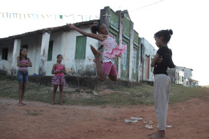 Foto com quatro crianças brincado, três estão brincando de elástico e uma está observando.