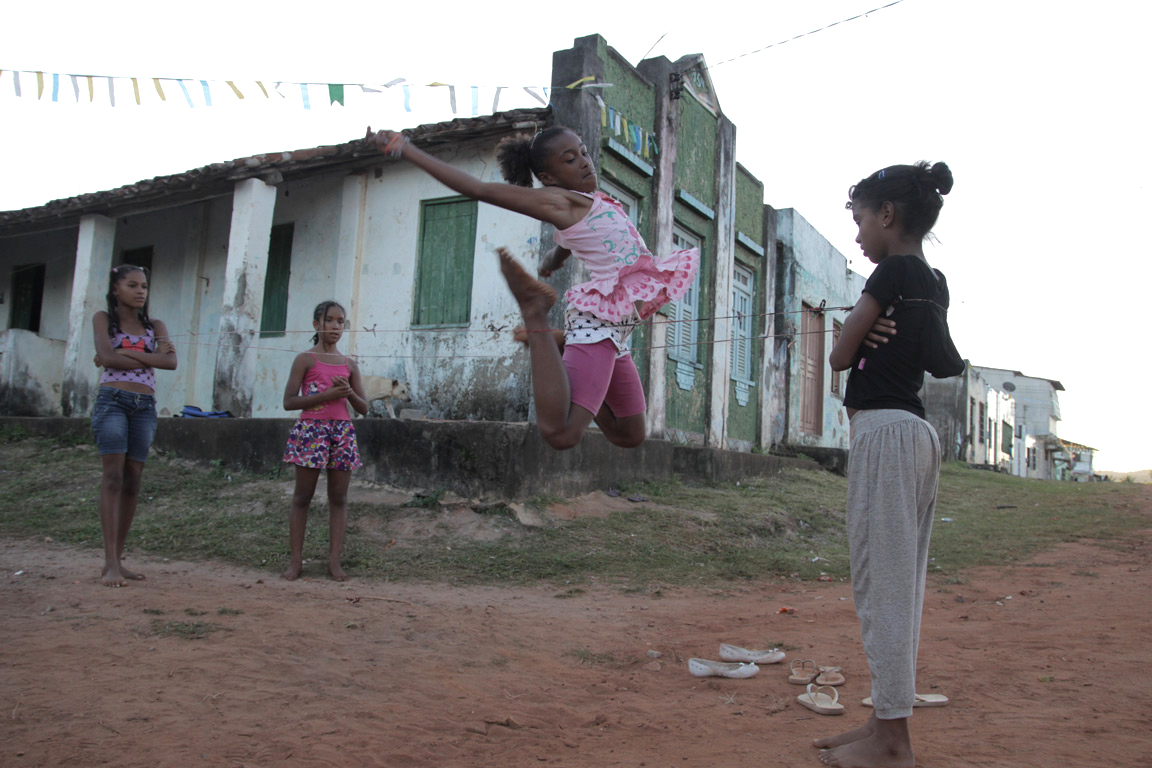 Foto com quatro crianças brincado, três estão brincando de elástico e uma está observando.