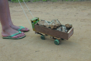 Foto de um caminhão de brinquedo feito de madeira carregando pedras em sua carreta, um garoto puxa o caminhão com uma linha de barbante.