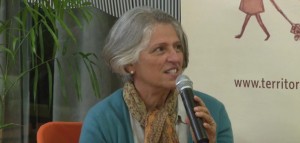 Foto de uma senhora grisalha sentada fala ao microfone.