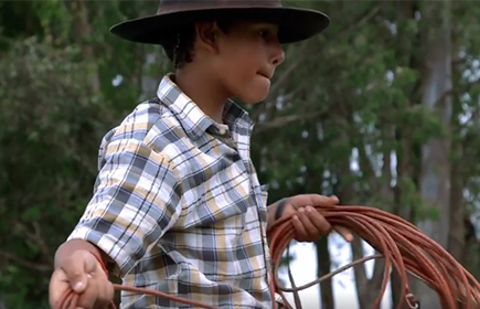 Foto de um garoto com chapéu marrom está girando um corda.