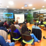 Foto de várias pessoas sentadas em círculo observam, uma mulher esta falando ao microfone.