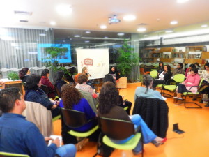 Foto de várias pessoas sentadas em círculo observam, uma mulher esta falando ao microfone.