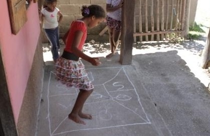 Foto com três meninas brincando, enquanto duas observam uma pula amarelinha.