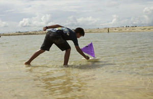 Foto de um garoto em um ribeirão, brinca com um barco a vela de brinquedo.