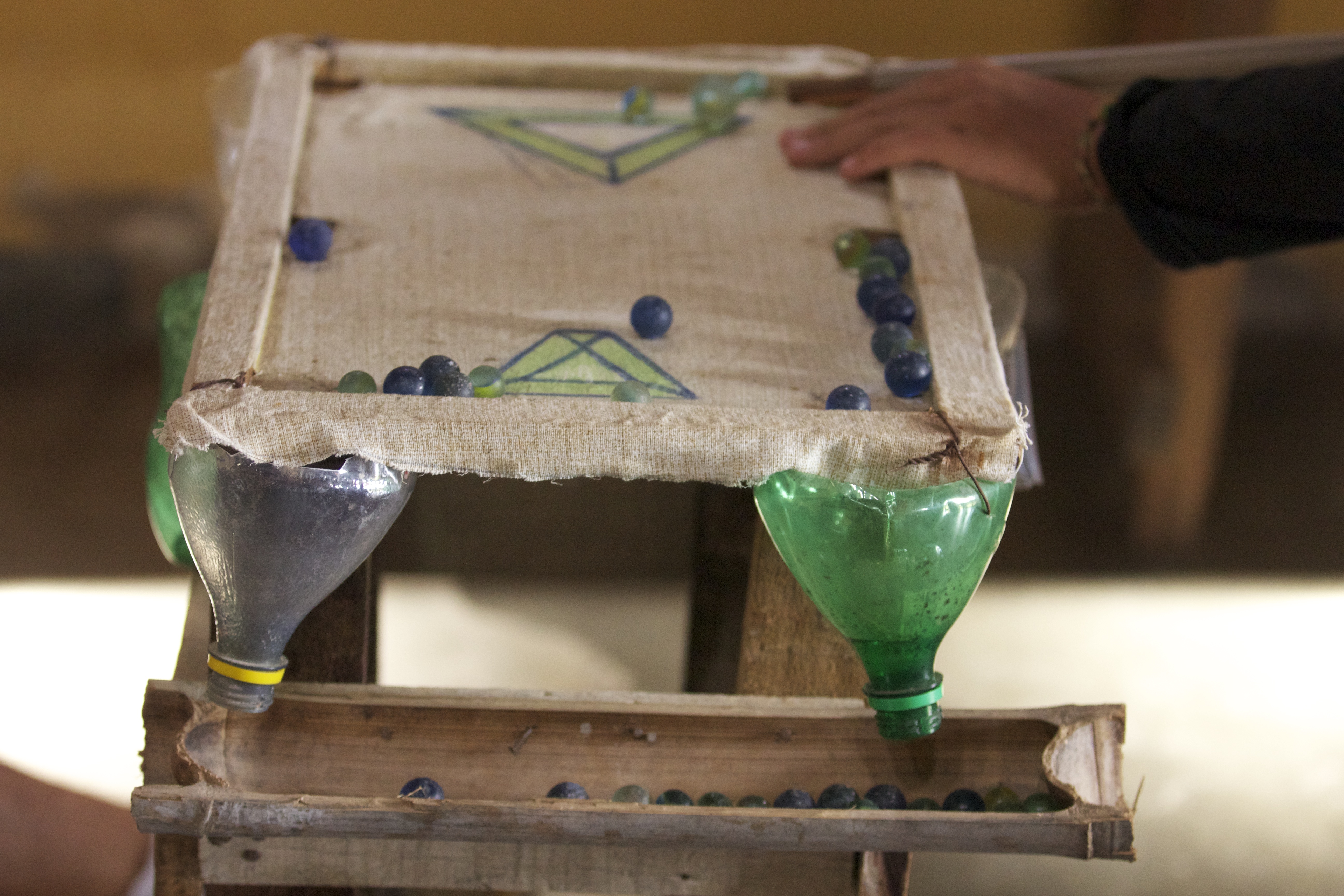 Foto de um brinquedo, mesa de sinuca feita com madeira, bambu e garrafa de plástico, bolinhas de gude estão nela.