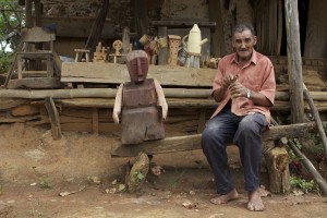 Foto de um senhor sentado em um banco de madeira feito a mão, ao lado direito dele há uma escultura de madeira feita artesanalmente.