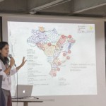 Foto com uma mulher falando ao microfone em cima de um palco, ao fundo um telão demonstra um gráfico de um mapa.