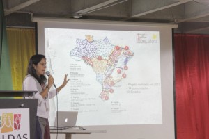 Foto com uma mulher falando ao microfone em cima de um palco, ao fundo um telão demonstra um gráfico de um mapa.