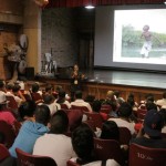 Foto de um salão com várias pessoas sentadas observando um homem sentado a beira de um palco falando ao microfone.