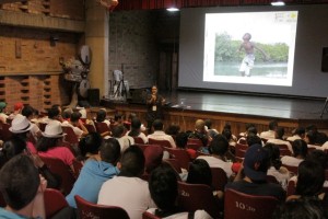 Foto de um salão com várias pessoas sentadas observando um homem sentado a beira de um palco falando ao microfone.