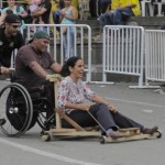 Foto composta por três pessoas, um homem de boné preto e óculos escuro empurra um cadeirante, o cadeirante de boné verde empurra uma mulher que está sentada em um carrinho de rolimã.