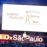 Foto de um salão com várias pessoas sentadas observando uma mulher interagindo, em cima de um palco.