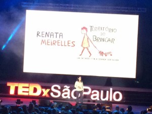 Foto de um salão com várias pessoas sentadas observando uma mulher interagindo, em cima de um palco.