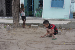 Foto onde uma criança brinca com uma tampinha de garrafa no chão de terra, enquanto outra observa apoiada em uma arvore usa uma máscara e segura um graveto de madeira.