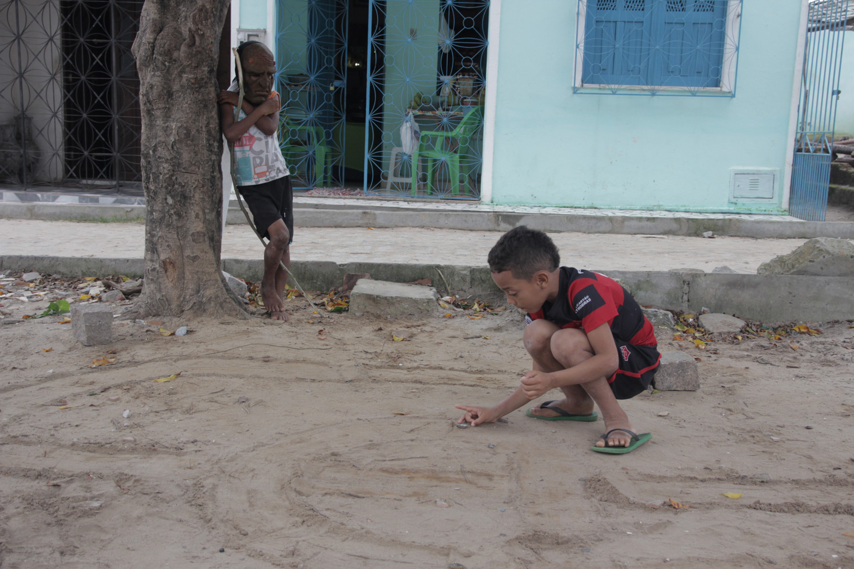 Foto onde uma criança brinca com uma tampinha de garrafa no chão de terra, enquanto outra observa apoiada em uma arvore usa uma máscara e segura um graveto de madeira.