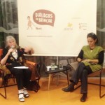 Foto com duas mulheres sentadas, uma senhora fala ao microfone enquanto gesticula com a mão esquerda, a sua esquerda uma outra mulher de pernas cruzadas escreve anotações em um caderno.