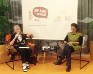 Foto com duas mulheres sentadas, uma senhora fala ao microfone enquanto gesticula com a mão esquerda, a sua esquerda uma outra mulher de pernas cruzadas escreve anotações em um caderno.