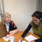 Foto com duas pessoas sentadas em uma mesa, uma senhora grisalha observa outra mulher que está sorrindo.
