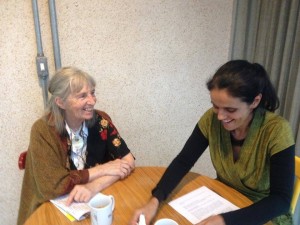 Foto com duas pessoas sentadas em uma mesa, uma senhora grisalha observa outra mulher que está sorrindo.