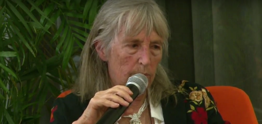 Foto de uma idosa grisalha falando ao microfone.