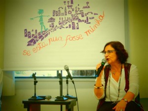 Foto de uma senhora sentada em uma cadeira, fala ao microfone.