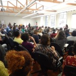 Foto composta por varias pessoas sentadas, observarão uma mulher em um palco falando ao microfone.