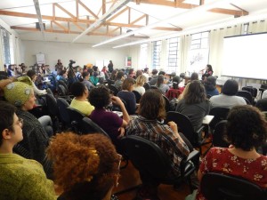 Foto composta por varias pessoas sentadas, observarão uma mulher em um palco falando ao microfone.
