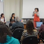 Foto composta por várias pessoas observando uma mulher falar ao microfone, a desta mulher duas outras mulheres observam.