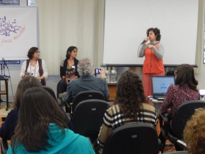Foto composta por várias pessoas observando uma mulher falar ao microfone, a desta mulher duas outras mulheres observam.