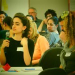 Foto composta por várias pessoas sentadas em mesas, em uma das mesas uma mulher fala ao microfone.