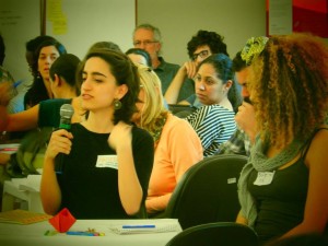 Foto composta por várias pessoas sentadas em mesas, em uma das mesas uma mulher fala ao microfone.