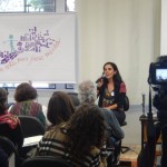 Foto composta por várias pessoas sentadas que observa uma mulher falar ao microfone em cima do palco, uma câmera está filmando.